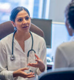 A young female mixed race doctor talks with her African patient about her medical concerns.  She is seated in her office at her desk and is wearing a white lab coat with a stethoscope around her neck.  She is using her hands to help articulate her point to the patient.  The patient is seated in front of the doctor and has her back to the camera.